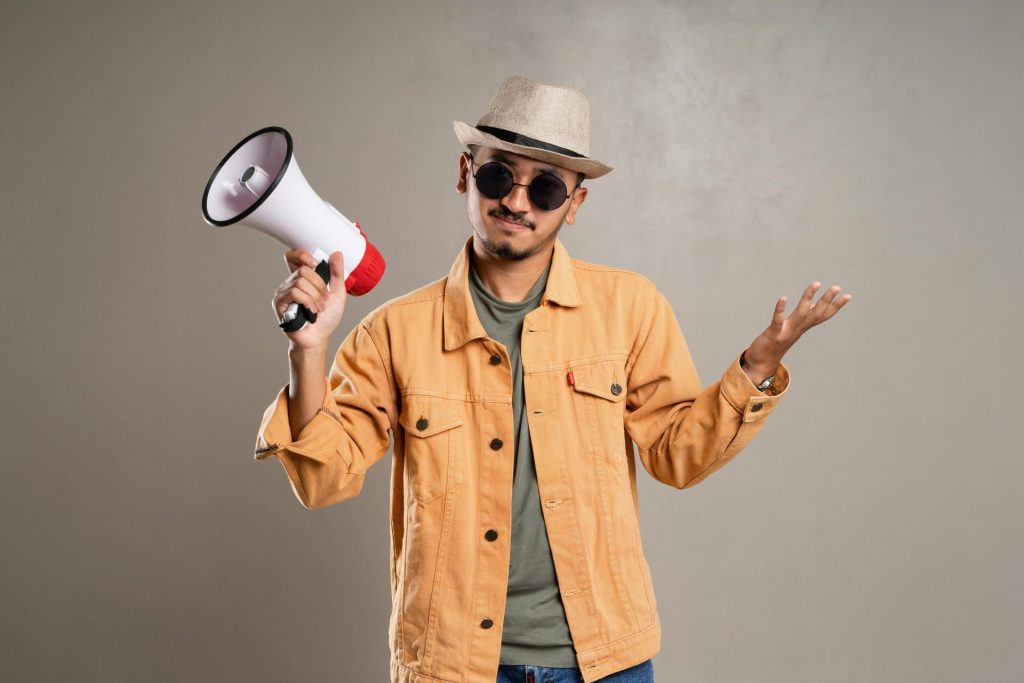 A fashionable man in sunglasses and a hat holding a megaphone with a confident stance, wearing a stylish jacket.